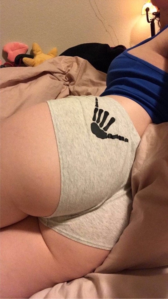 another picture of clarke laying on a dusty rose duvet, with his body twisted at an angle to display his ass. you can see a few of her stuffies, a black sweatshirt, as well as a lamp in the background.