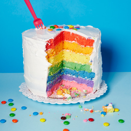 [ID: picture of a cake with white frosting on the outside, and rainbow layers on the inside | a big 