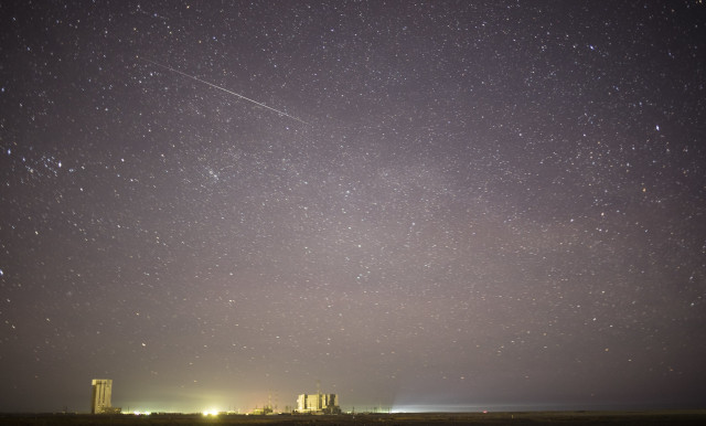 In this long exposure, a meteor streaks across a dusty blue star-spangled sky. Along the horizon, the bright lights of the Baikonur Cosmodrome glow yellow, illuminating buildings and a launch pad. Credit: NASA/Joel Kowsky