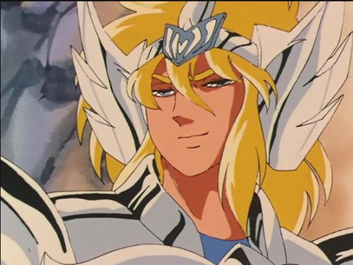 An image of one of the knights, Cygnus, cropped close to his face. He has fluffy blonde hair and blue eyes. His armor headpiece is stylized as a swan. His entire outfit is very very shiny and he is smiling at someone out of frame.