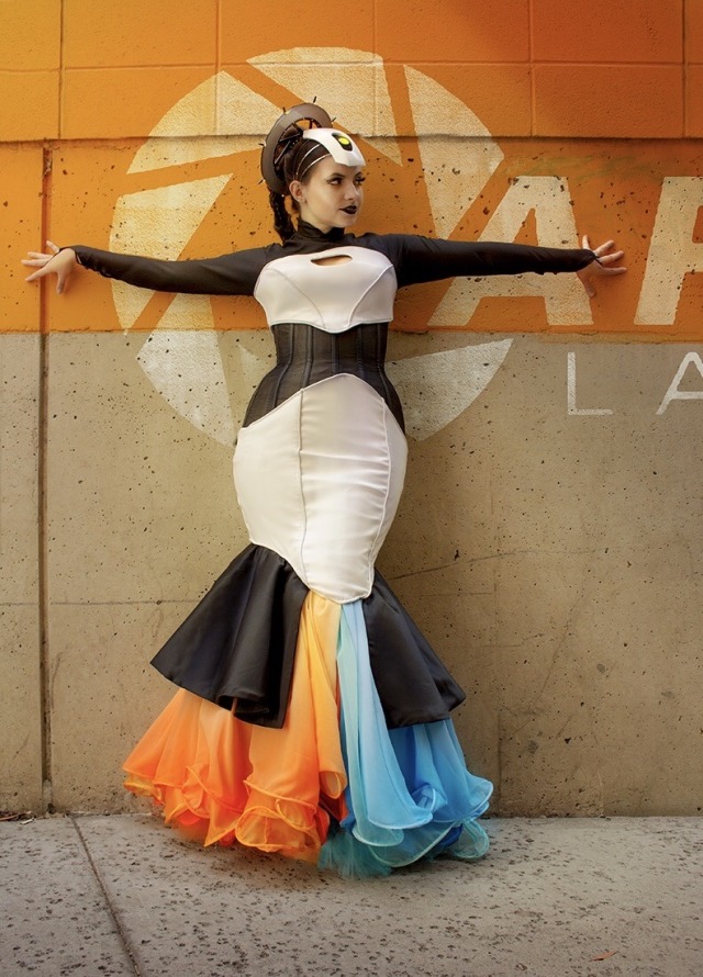 a woman wearing a gown inspired by the portal game series and the character glados. the dress is corseted, long-sleeved, and mermaid-shaped. it is black and white with a blue and orange skirt.