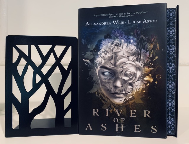 Shown is the Twisted Retreat special edition of RIVER OF ASHES (with cover art depicting a face merging into flowers and sprayed edges with skulls) sitting on a white shelf beside a black bookend in the shape of a stylized woods. Photo by AHS.