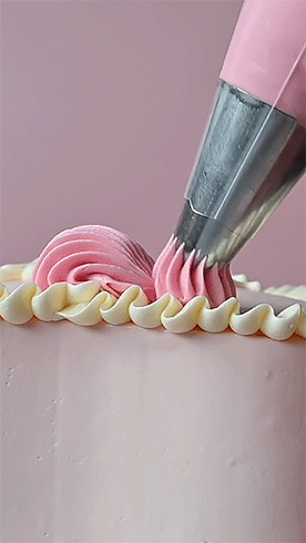 light pink frosting being piped in ruffles on top of cake that already has a thinner layer of white decorative frosting