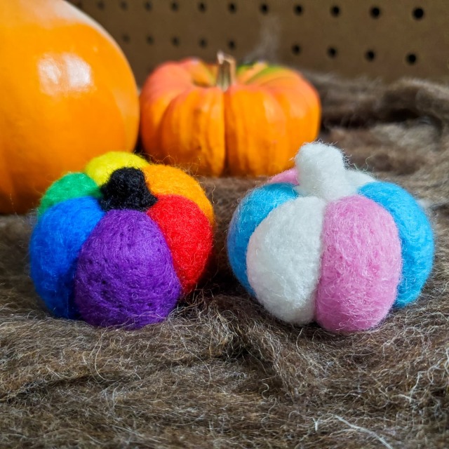 a photo of two needle felted pumpkins and two real pumpkins on top of brown wool. The felted pumpkins are colored to look like the rainbow pride flag and the transgender flag.