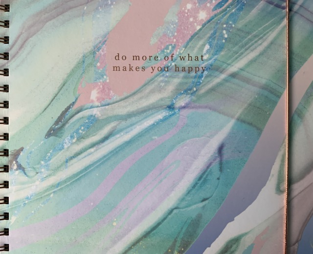 Cropped, close-up image of a ring-bound scrapbook, with a blue, purple and pink marble effect design on the front cover. At the top is text in rose gold lettering which reads: "do more of what makes you happy".