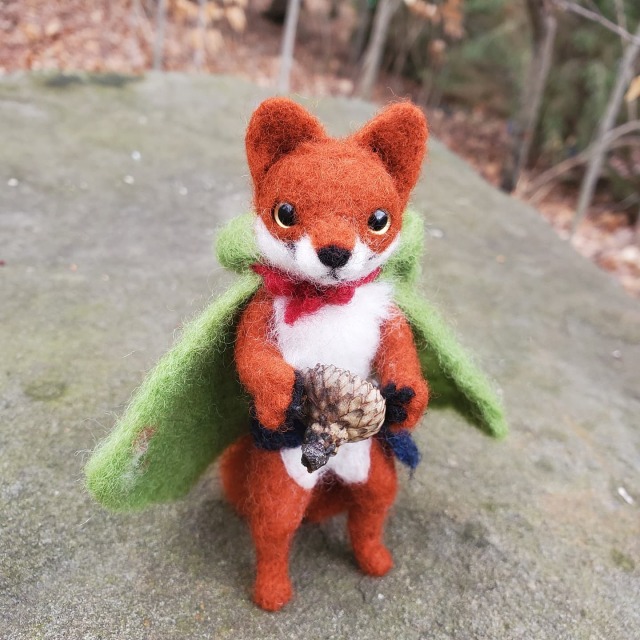 A photo of a needle felted bipedal fox with a green cape and blue belt in the forest. They are standing on a rock and are holding an acorn. Their hood is down, showing their ears