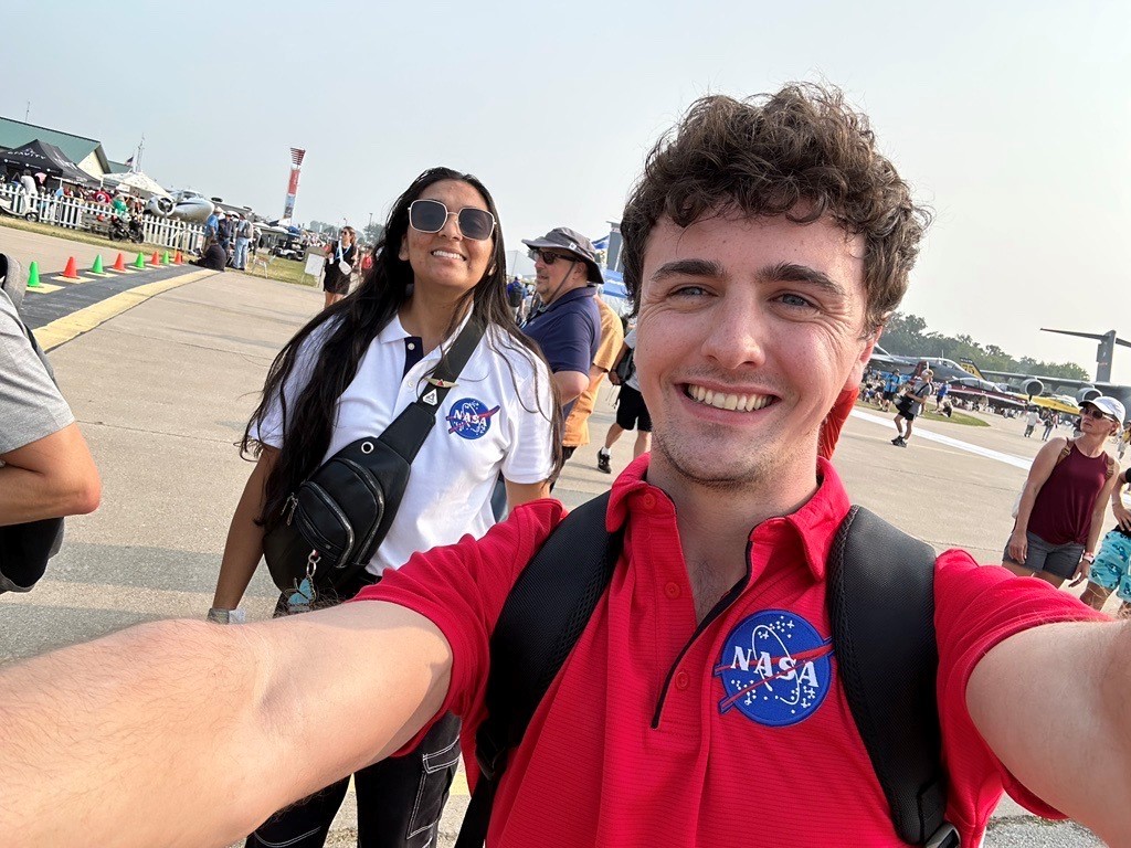 Two people pose for a photo in a street. The person taking the photo is taking the image “selfie style,” so that their arms are visible in the frame. Both of the people are smiling. One is wearing a white polo and the other is wearing a red polo, and both shirts feature the NASA insignia. People can be seen milling about behind the two who are posing for the photo, and in the distance, small aircraft parked on grass can also be seen. Credit: NASA