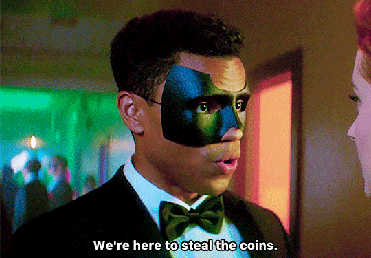 GIF FROM EPISODE 1X06 OF NANCY DREW. CLOSE-UP OF NICK AT THE VELVET MASQUE PARTY, WEARING A MASK THAT COVERS THE TOP HALF OF HIS FACE. THERE ARE PARTY-GOERS IN THE BACKGROUND. NICK IS TALKING TO NANCY WHO IS JUST BARELY IN FRAME. NICK SAYS "WE'RE HERE TO STEAL THE COINS."
