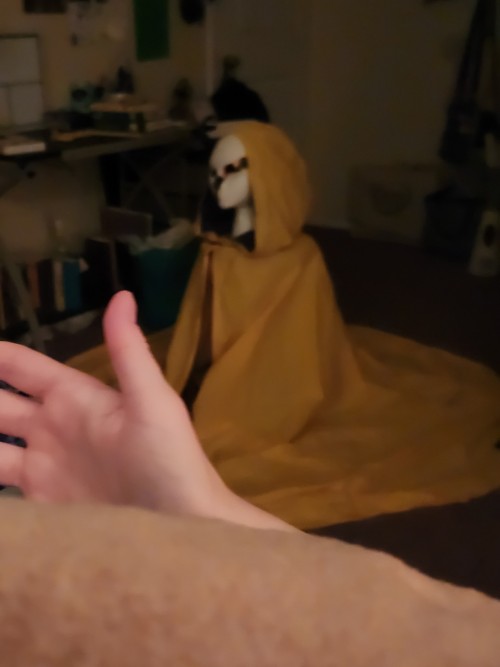 A blurry picture of the cloaked figure on the floor of a bedroom, with a hand in the foreground gesturing loosely to it. The light is dimmer and the figure looks more like a solid figure than a cloak on a stool.