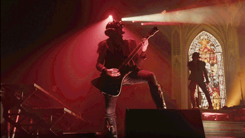 Phantom head-bangs while playing guitar. His foot is up on a stage monitor. Swiss ghoul dances behind him.