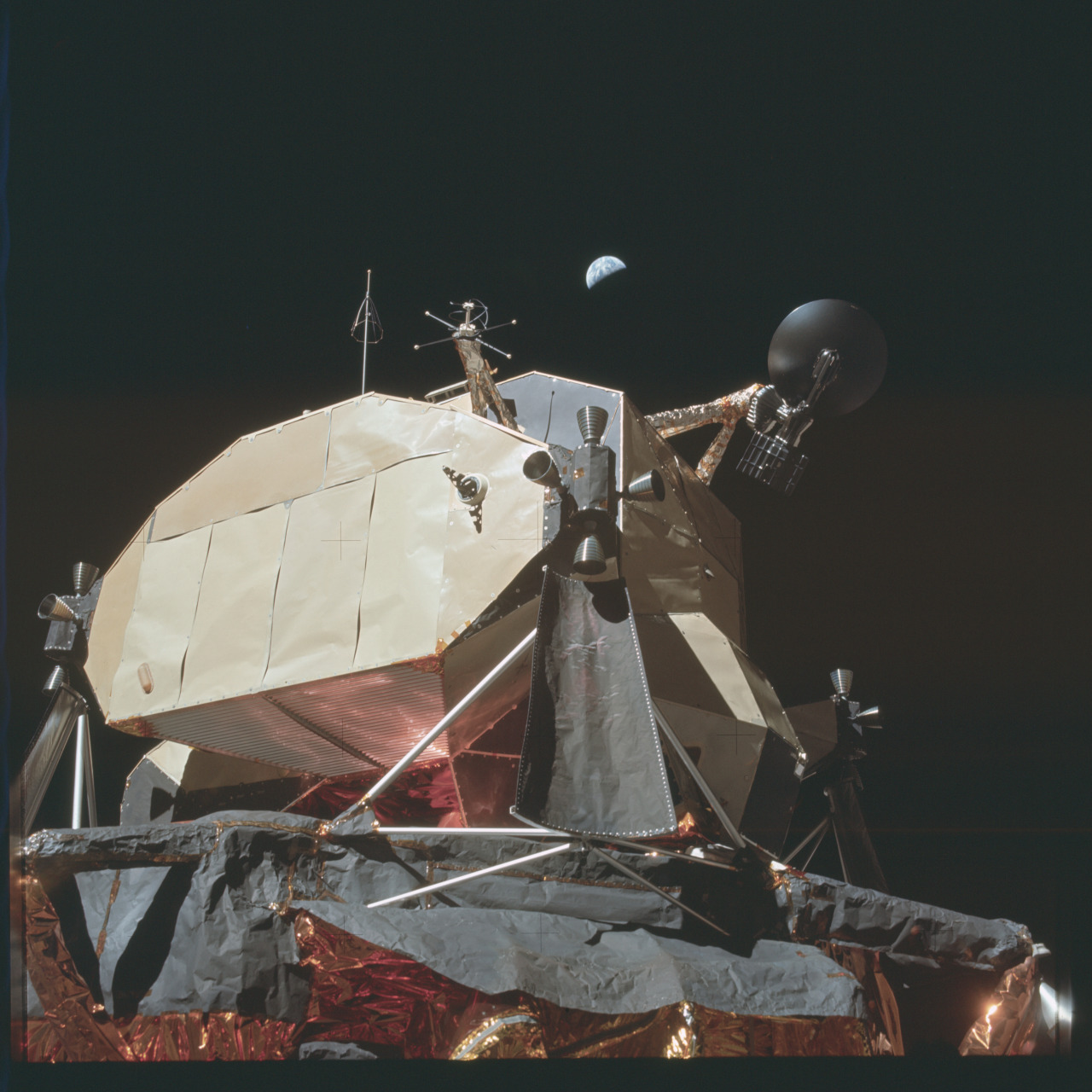 The half Earth appears in the black sy over the Lunar Module on the lunar surface. The spacecraft has a radio dish, black thermal blankets, and a tubular metal support structure. Credit: NASA