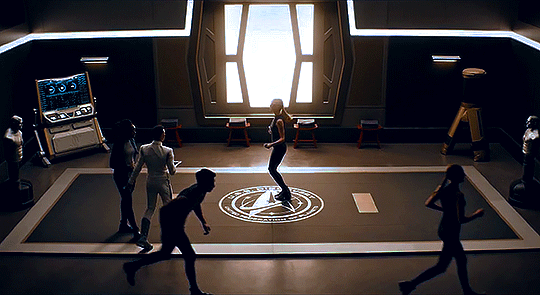 Wide shot of the gym on the USS Discovery, with a large Starfleet insignia in the centre where Detmer continues jumping rope. 