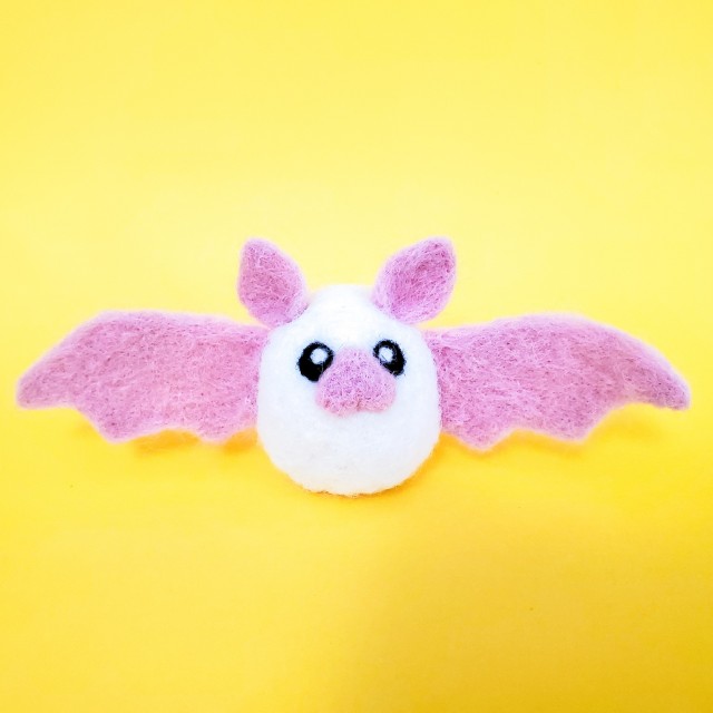 A photo of a white needle felted bat with a pink nose, ears, and wings. It is sitting on a yellow background.