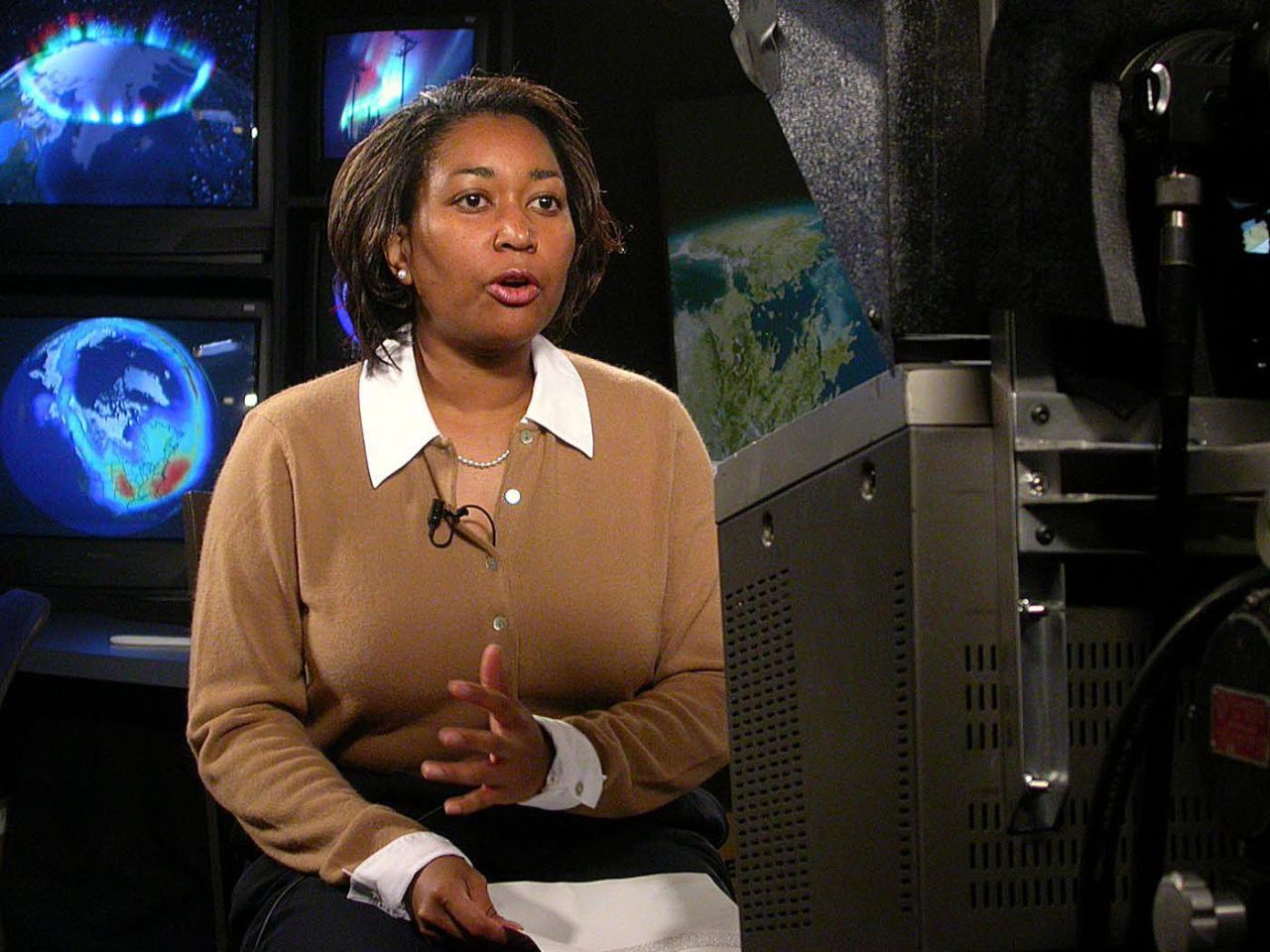 Black woman sitting in front of a camera that is slightly off-frame. She is wearing a brown sweater with a white collared shirt underneath. There are images of Earth from space behind her. Credit: NASA