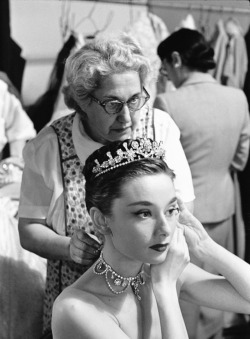 rareaudreyhepburn:  Audrey Hepburn (with Edith Head in the background) puts on her tiara and necklace while on the set of Roman Holiday, 1952.