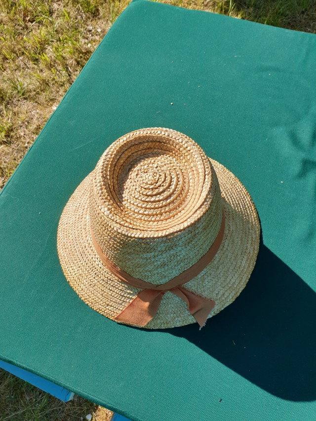 a straw hat with a dull orange ribbon lying on a green mattress. the brim is longer on one side and the crown had a trilby like indentation 