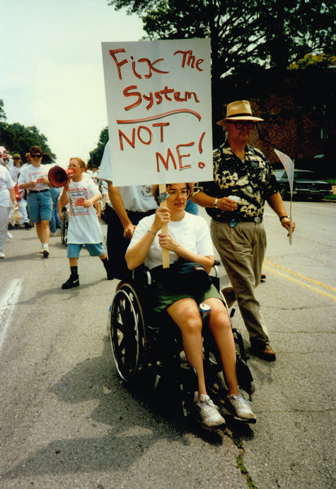 socialismartnature:A protester who uses a wheelchair holds up a sign that says “Fix The System: Not 