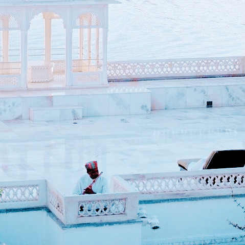 a shot of the taj lake palace resort in udaipur, india, a gorgeous marble structure facing a large lake. an indian man in a blue and pink turban plays a flute on the terrace. 