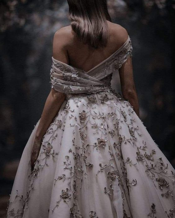 An image of a woman in a white and gold off-the-shoulder ballgown looking away