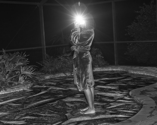 Black and white photograph by Widline Cadet of a figure whose face is obscured by a bright flash of light stands in a velvet robe on top of a pool surrounded by the darkness of night.