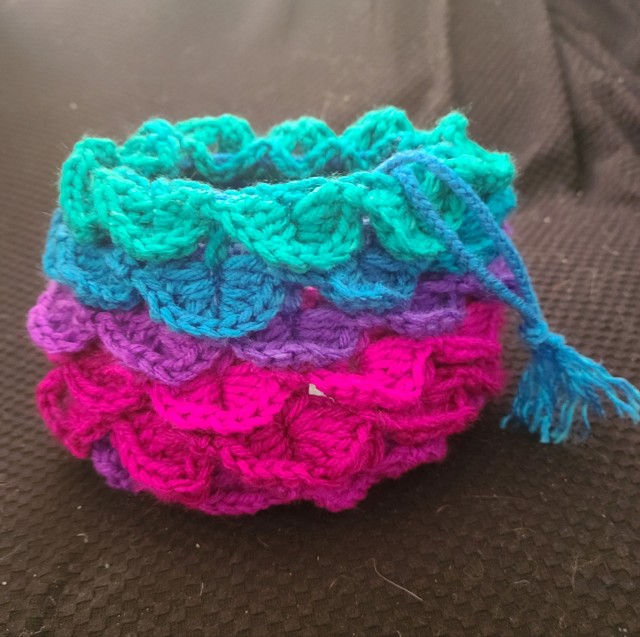 A crocheted bag using the crocodile stitch. Each row is a different colour, the bottom four are shades of purple and pink, the top two are blue and green