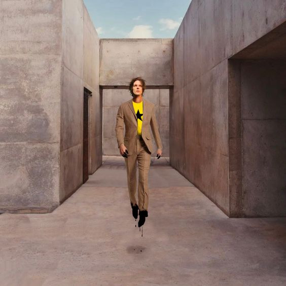 An image of Dallon Weekes hovering above the ground in some plain industrial concrete structure. His arms and legs are bent in such a way that implies he is walking. He is now wearing a brown suit and a shirt with a satr on it, and the same black ichor is dripping from his fingers and feet