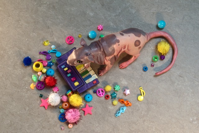 A photo of a hand-sized painted clay figurine of Neil the hairless rat, playing a small purple synthesizer. The photo is taken slightly from above, showing Neil\'s left side.  The knobs, wheels and black keys of the synth are in the colors of the rainbow. The figurine has its right forepaw raised, resting on the keys.  The figurine sits on a grey vinyl floor, and is surrounded by colorful glittery pompoms and plastic beads in a variety of shapes laid out around it. Some of the beads are shaped like stars and musical notes.