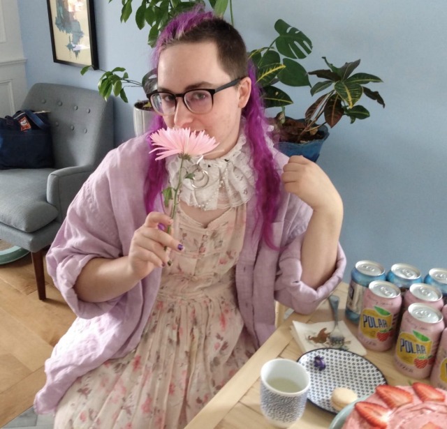 a nonbinary person with long purple hair holds a pink flower against their face. they're dressed in a pink floral dress with a light purple buttondown worn loose like a cardigan, and a lace ruff decorated with hardware and fake pearls 