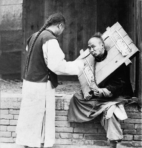 Chinese man feeding a criminal in a cangue with a sign describing his crime, 1905