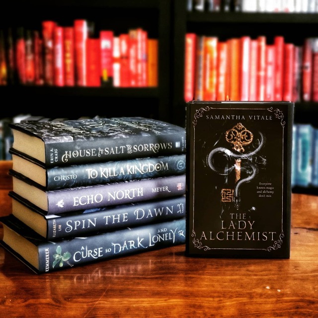 The Lady Alchemist beside a stack of YA fairy tale retellings: house of salt and sorrows, echo north, to kill a Kingdom, spin the dawn, and a curse so dark and lonely. bookshelf in background.
