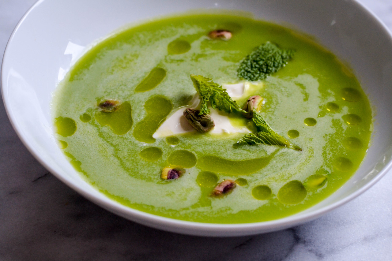 Image of a green pea soup in a white bowl with crème fraîche, pistachios, extra virgin olive oil, and mint leaves