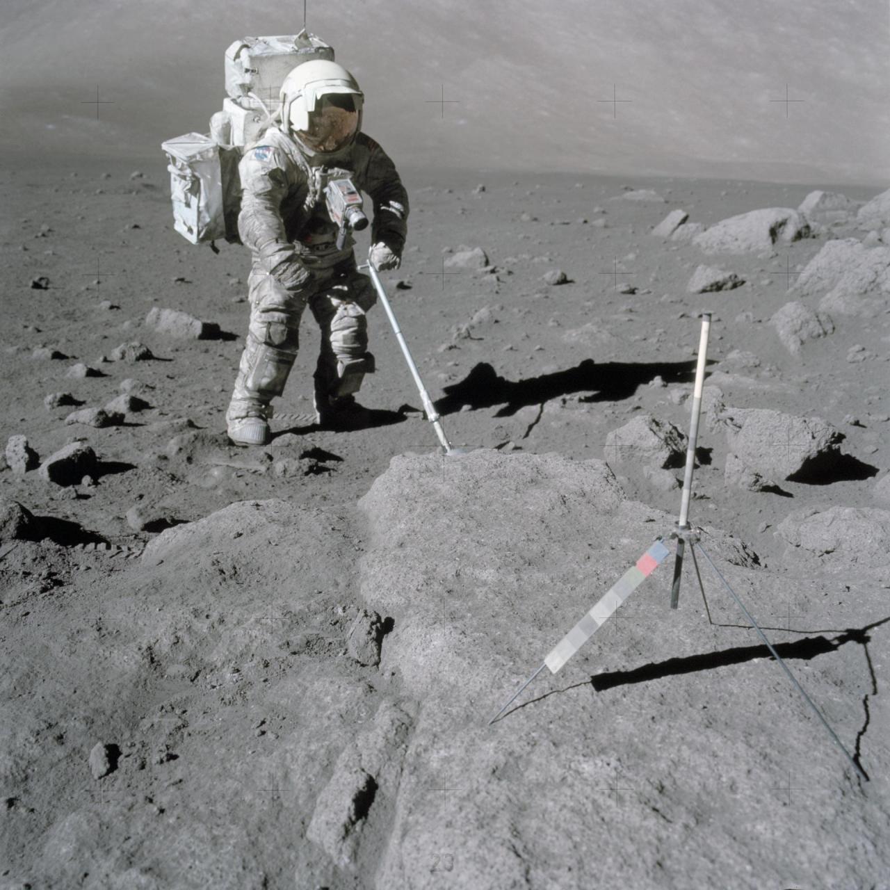 Scientist-astronaut Harrison Schmitt, Apollo 17 lunar module pilot, uses an adjustable sampling scoop to retrieve lunar samples during the second Apollo 17 extravehicular activity (EVA), at Station 5 at the Taurus-Littrow landing site. A gnomon is atop the large rock in the foreground. The gnomon is a stadia rod mounted on a tripod, and serves as an indicator of the gravitational vector and provides accurate vertical reference and calibrated length for determining size and position of objects in near-field photographs. The color scale of blue, orange and green is used to accurately determine color for photography. Credit: NASA