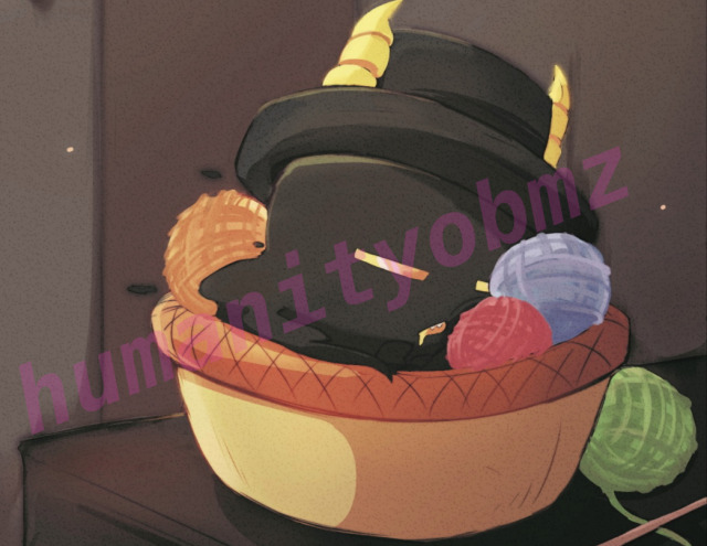 A little D design where he’s sleeping in a yarn basket with three yarn balls next to him. The background is a slightly out of focus wall. On the table is some knitting needles. Over top of the design is transparent pink text that reads humanityobmz.