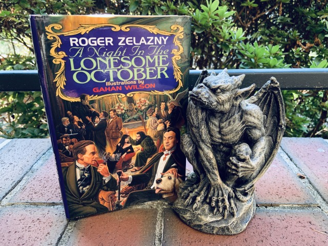 A picture of A NIGHT IN THE LONESOME OCTOBER (with cover art depicting a meeting of many familiar Gothic characters, including Sherlock Homes and Count Dracula in the forefront) sitting on a brick walk beside a stone gargoyle.