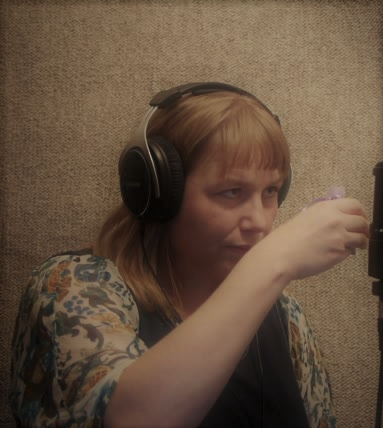 A warm photo of Katherine Seaton, wearing headphones and seated at the microphone, squeezing a lavender bag in one hand