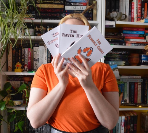 me, in front of my bookshelves, holding the three books of the all for the game trilogy in front of my face, covers showing to the camera