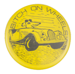 a yellow pin with a drawing of a dog driving a vintage car. black text on the border reads 'BITCH ON WHEELS'