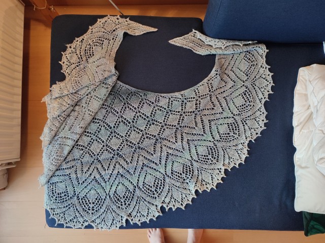 The same light blue handknit lace shawl, draped over a dark blue sofa for display. It is freshly washed and blocked, and its shape is far better defined and symmetrical and the stitch pattern very clear. It has grown a little bit.