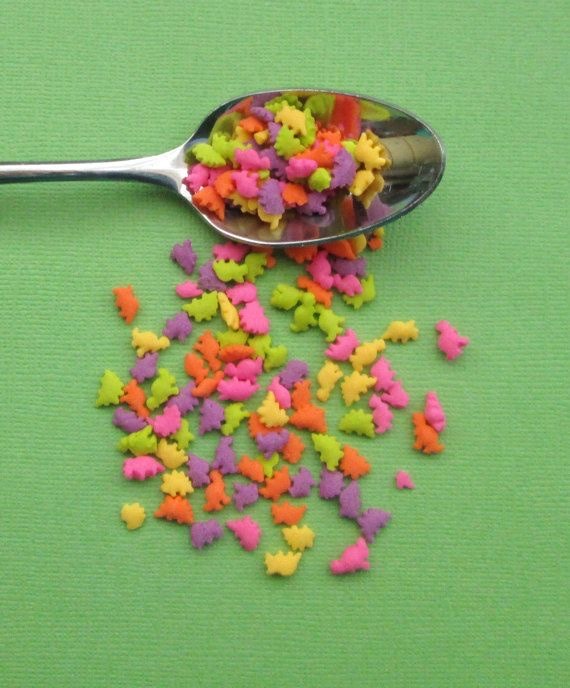 A spoon laid on a matte green background, spilling brightly coloured dinosaur-shaped sprinkles in pink, purple, green, yellow, and orange.