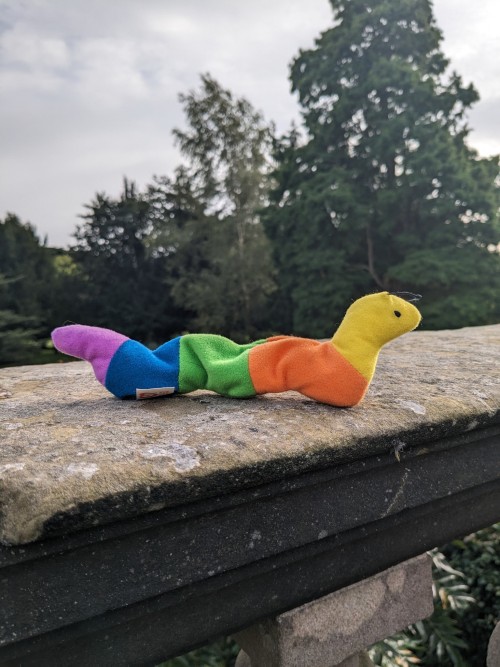 an inch worm beanie baby sat on a stone fence, there's trees in the background and the sky is scattered with clouds