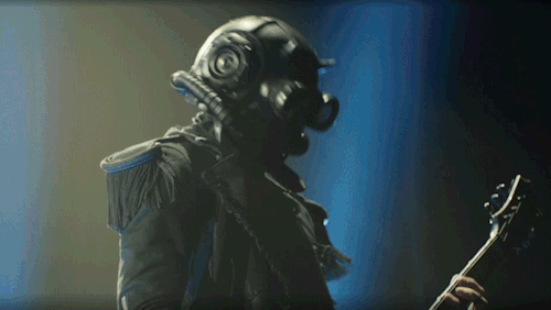 A close up of Phantom's chest. His masked head is barely visible as he turns toward the camera, lifting his strumming hand to pat his chest. His index and thumb still hold his pick, but the rest of his fingers are splayed out, showing the black tape around his ring finger. 