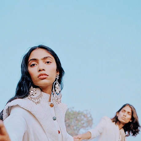 two south asian people wearing white clothes and jewellery stand against a clear blue sky. one of them reaches out to the camera, the other, indistinctly visible, looks towards their companion.