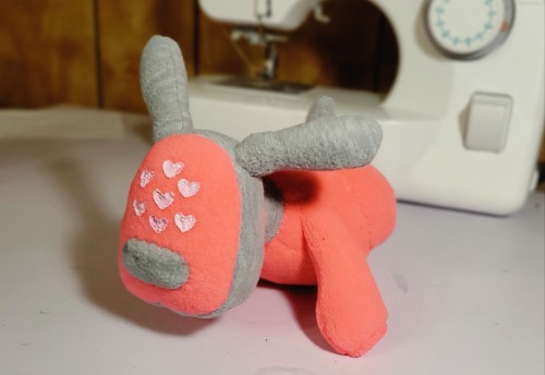 A photograph of a plush that is made to resemble an iDog. This one is pink and facing the camera, with pink hearts on the face.