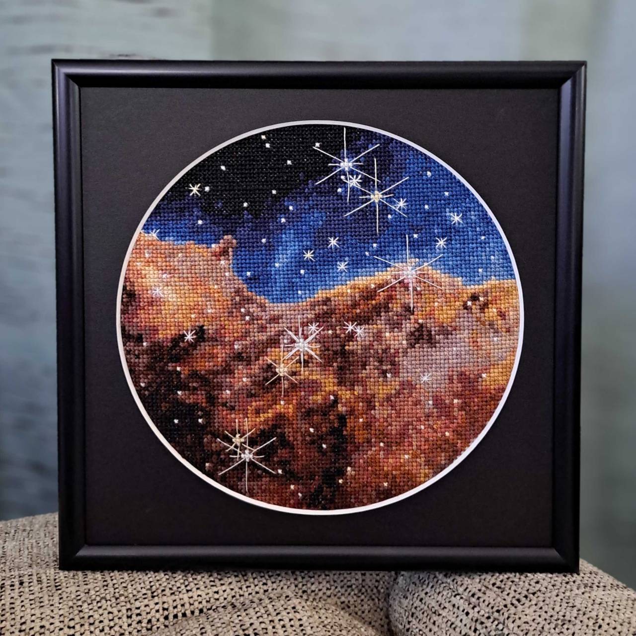 hThis embroidered image shows the Carina Nebula captured by the James Webb Space Telescope. The image is framed in black. At the center a circular piece of art appears outlined in white. At the top of the circle, the thread is dark blue on the left. As you travel down white stars appear in lighter shades of blue. In the middle threads turn to dark black, red and orange to signify the nebula’s gas-like structure.