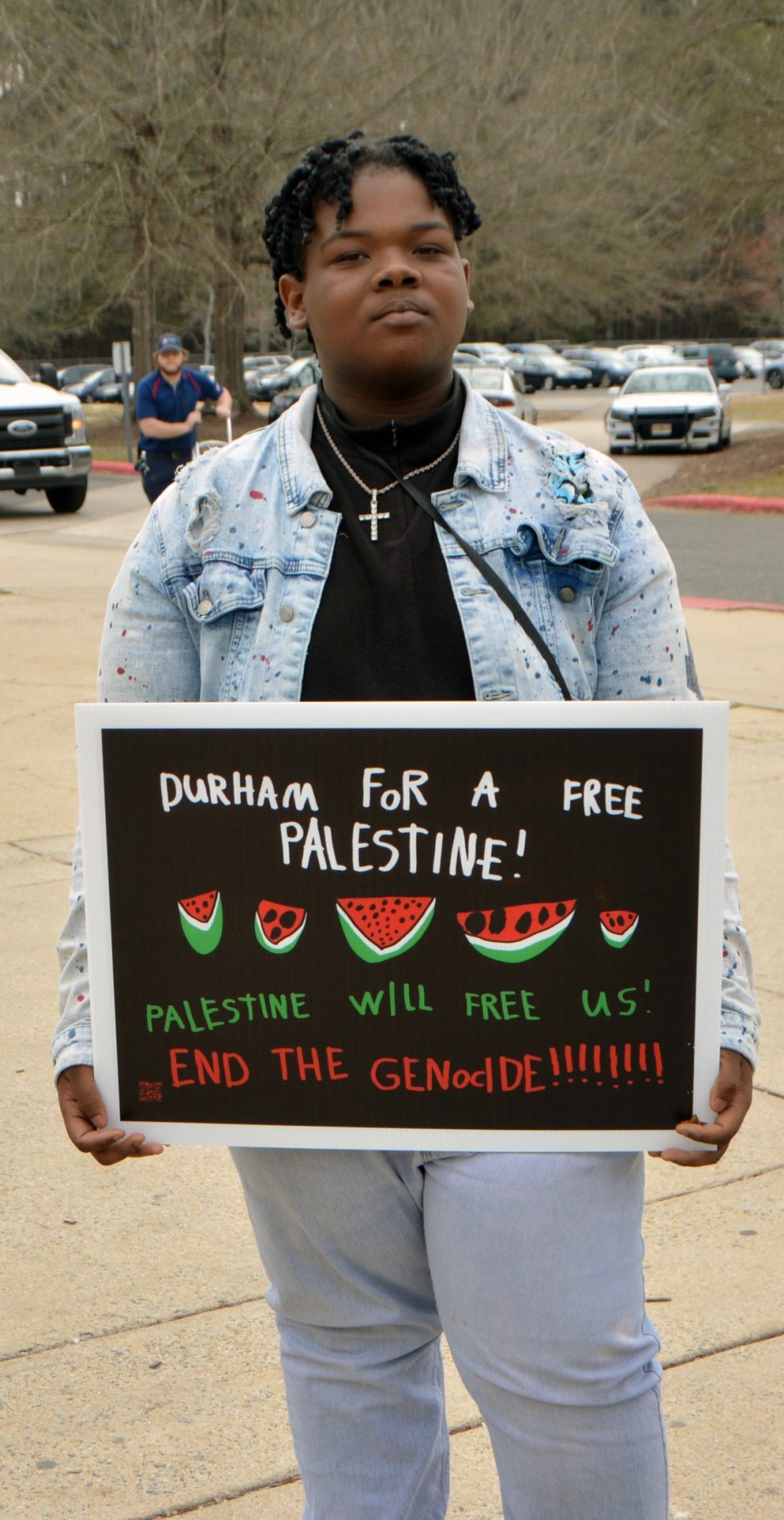 A photo of a young Black high school student standing with a placard. The placard reads, “Durham for a free Palestine! Palestine will free us! End the genocide!” and has drawings of five slices of watermelon in the center.