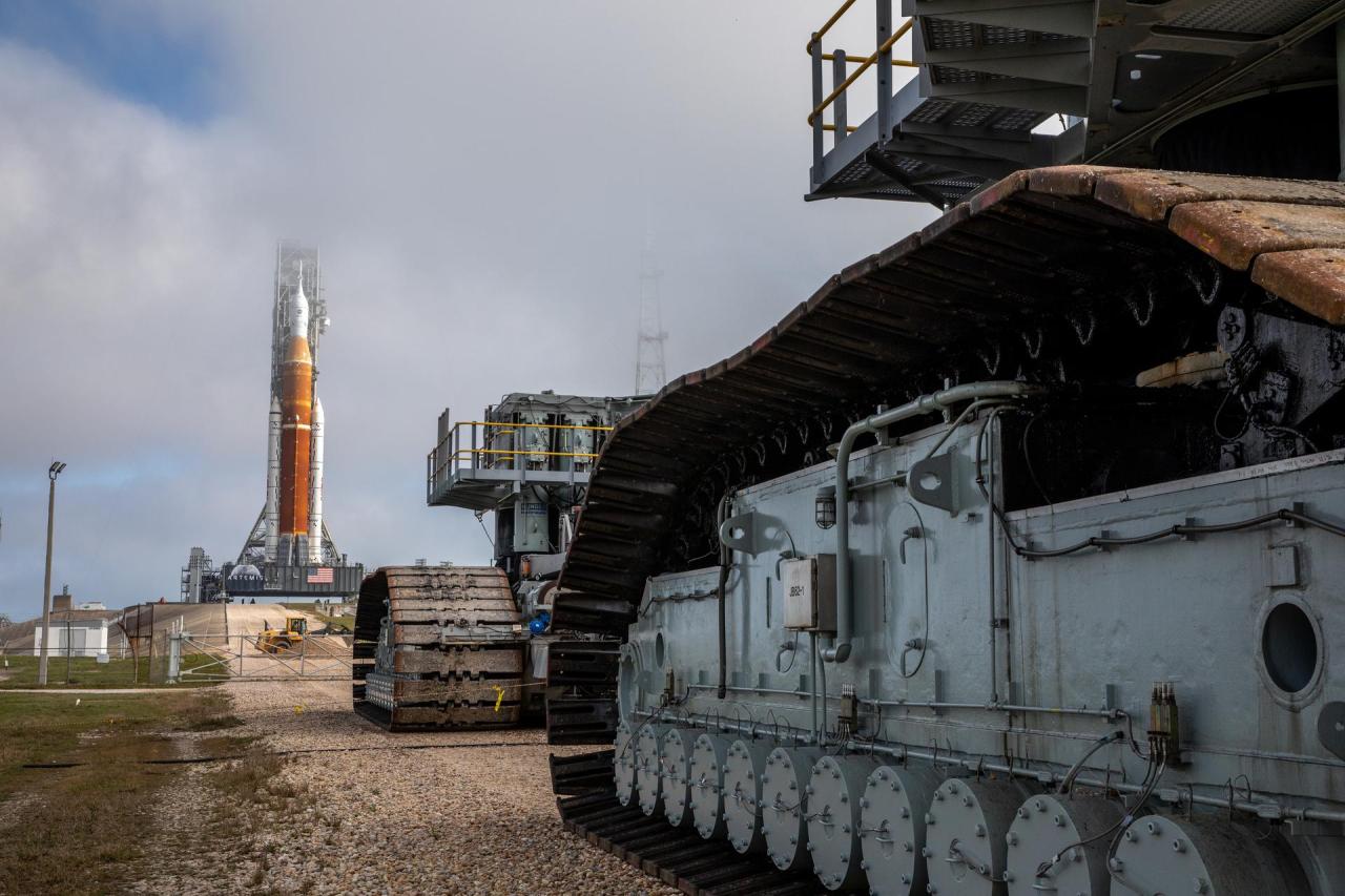 The Space Launch System rocket stands upright on the launchpad. The background is the sky dominated by clouds. The rocket has an orange central fuel tank with two white rocket boosters on either side. The Crawler-Transporter 2 is in the foreground with its massive tread-like wheels. Credit: NASA/Kim Shiflett