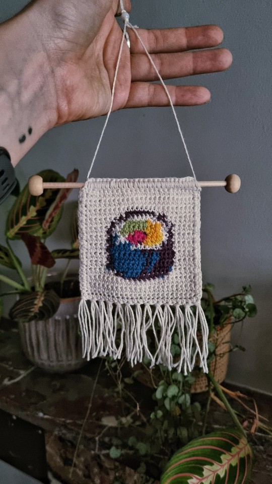 A hand holding a crochet tapestry depicting a sushi roll.