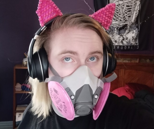 valentine, a white nonbinary person with blonde side shaved hair and bright blue eyes, staring into the camera, wearing a gray respirator with pink p100 filters and a pair of big black bulky headphones with magenta kandi cat ears attached