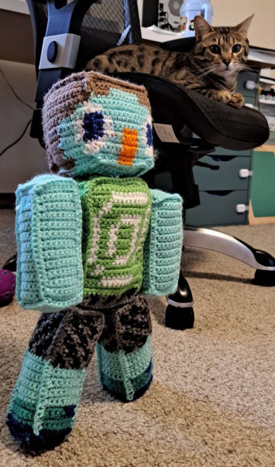Image 4: a photo of the juppet doll, standing upright. The photo is taken from an angle so you can see a spotted tabby cat sitting on a chair in the background, looking at the camera.
