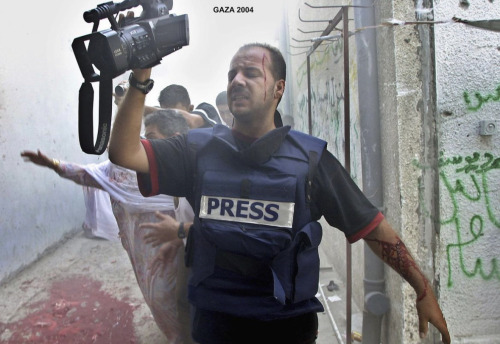 A journalist wearing a press jacket and holding a camera over his head. His head is injured and bleeding. One of his arms is bleeding badly. Behind him, there's blood on the floor and people running. There is text on the image that says 'Gaza 2004'